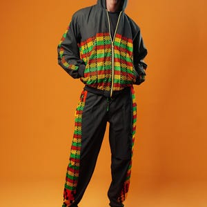 Man standing against an orange background wearing black tracksuit trousers with colourful Kente African print panels down the sides. The trousers have a relaxed fit with elastic cuffs at the ankles and a drawstring waist.