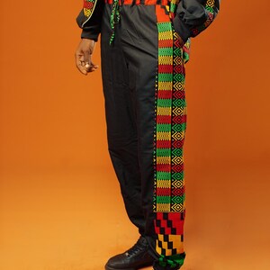 Man standing against an orange background wearing black tracksuit trousers with colourful Kente African print panels down the sides. The trousers have a relaxed fit with elastic cuffs at the ankles and a drawstring waist.