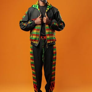 Man standing against an orange background wearing black tracksuit trousers with colourful Kente African print panels down the sides. The trousers have a relaxed fit with elastic cuffs at the ankles and a drawstring waist.