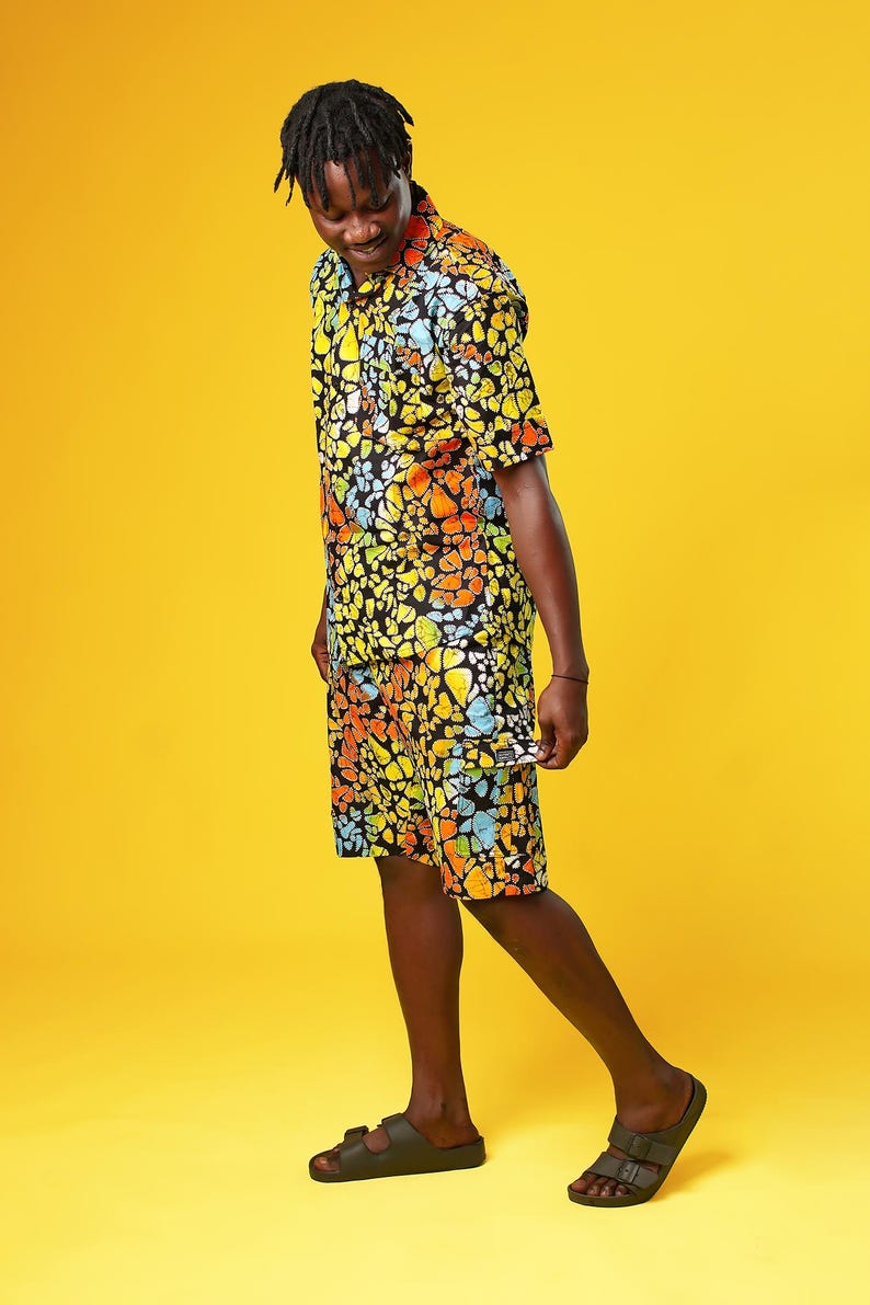 A man stands in front of a yellow background wearing Funky Men’s Cargo Shorts made from multicoloured African print fabric with a butterfly pattern in orange, yellow, blue, and black. The shorts have a relaxed fit and side pockets, paired with a matching short-sleeve shirt. This is a bold Summer Hawaiian Shorts look, ideal for African Print Men’s Clothing, a Hippie Festival Set, or a Carnival Outfit from Ashanti Empress.