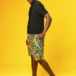 A man stands in front of a yellow background wearing Funky Men’s Cargo Shorts made from multicoloured African print fabric with a butterfly pattern in orange, yellow, blue, and black. The shorts have a relaxed fit and side pockets, paired with a matching short-sleeve shirt. This is a bold Summer Hawaiian Shorts look, ideal for African Print Men’s Clothing, a Hippie Festival Set, or a Carnival Outfit from Ashanti Empress.