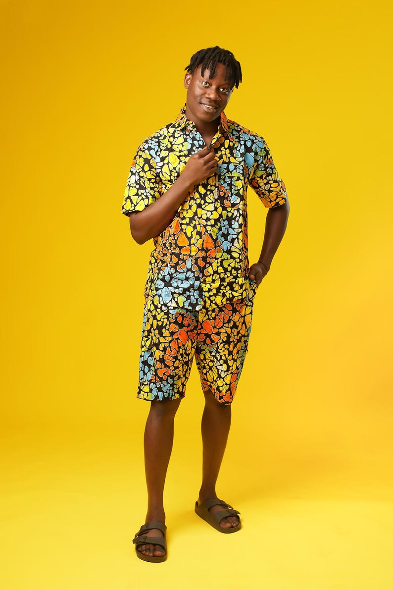 A man stands in front of a yellow background wearing Funky Men’s Cargo Shorts made from multicoloured African print fabric with a butterfly pattern in orange, yellow, blue, and black. The shorts have a relaxed fit and side pockets, paired with a matching short-sleeve shirt. This is a bold Summer Hawaiian Shorts look, ideal for African Print Men’s Clothing, a Hippie Festival Set, or a Carnival Outfit from Ashanti Empress.