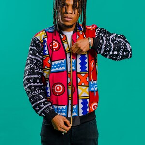 May include: A man wearing a colorful patterned bomber jacket with black and white geometric designs on the sleeves. The jacket has a zipper closure and is unzipped. The man is wearing black jeans and has dreadlocks.