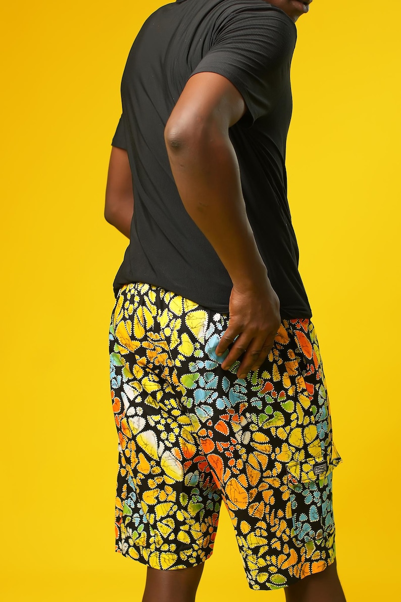 A man stands in front of a yellow background wearing Funky Men’s Cargo Shorts made from multicoloured African print fabric with a butterfly pattern in orange, yellow, blue, and black. The shorts have a relaxed fit and side pockets, paired with a matching short-sleeve shirt. This is a bold Summer Hawaiian Shorts look, ideal for African Print Men’s Clothing, a Hippie Festival Set, or a Carnival Outfit from Ashanti Empress.