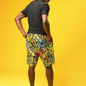A man stands in front of a yellow background wearing Funky Men’s Cargo Shorts made from multicoloured African print fabric with a butterfly pattern in orange, yellow, blue, and black. The shorts have a relaxed fit and side pockets, paired with a matching short-sleeve shirt. This is a bold Summer Hawaiian Shorts look, ideal for African Print Men’s Clothing, a Hippie Festival Set, or a Carnival Outfit from Ashanti Empress.