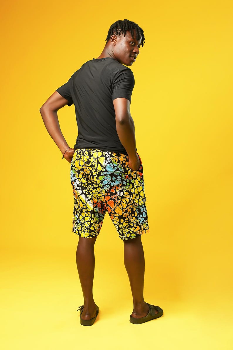 A man stands in front of a yellow background wearing Funky Men’s Cargo Shorts made from multicoloured African print fabric with a butterfly pattern in orange, yellow, blue, and black. The shorts have a relaxed fit and side pockets, paired with a matching short-sleeve shirt. This is a bold Summer Hawaiian Shorts look, ideal for African Print Men’s Clothing, a Hippie Festival Set, or a Carnival Outfit from Ashanti Empress.