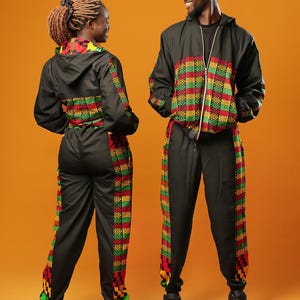 Woman and man standing against an orange background wearing black tracksuit trousers with colourful Kente African print panels down the sides. The trousers have a relaxed fit with elastic cuffs at the ankles and a drawstring waist. They both pair them with matching black jackets featuring Kente trim.