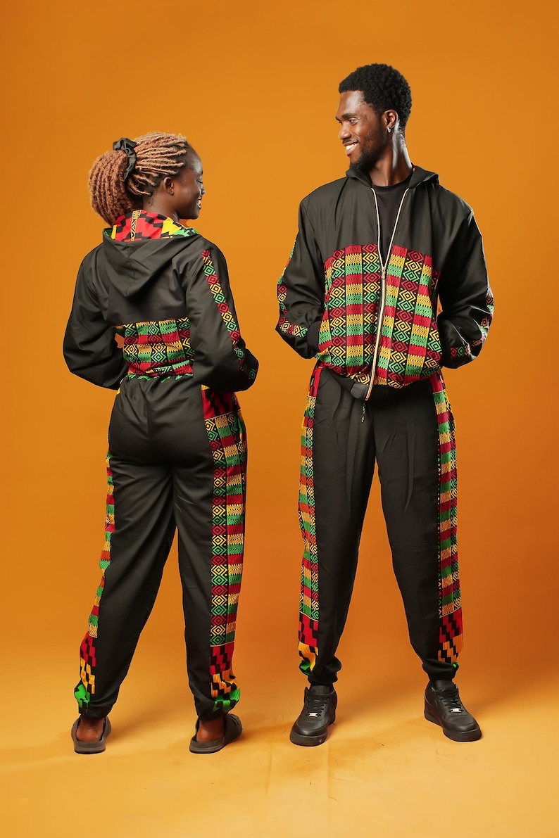 Woman and man standing against an orange background wearing black tracksuit trousers with colourful Kente African print panels down the sides. The trousers have a relaxed fit with elastic cuffs at the ankles and a drawstring waist. They both pair them with matching black jackets featuring Kente trim.