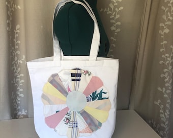 May include: White tote bag with a large, colorful flower applique. The flower design features various fabric patterns in pastel shades of pink, yellow, and blue. The bag has white handles and is displayed against a neutral background.