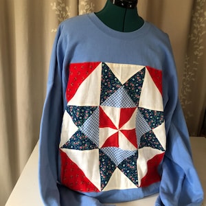 May include: A light blue sweatshirt featuring a patchwork star design. The star is composed of red, white, blue floral, and gingham patterned fabric. The sweatshirt has long sleeves and a crew neck.