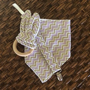 May include: A baby pacifier holder with a wooden ring and a fabric section featuring a chevron pattern in lavender, olive green, and white. The holder has a metal clip and a white ribbon.