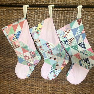 May include: Three Christmas stockings with patchwork patterns in various colors, including red, blue, green, and pink. Each stocking has a solid pink toe and lining, and is suspended by a white clip.