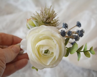 Puede incluir: Un prendedor floral con una gran ranúnculo blanca, un cardo verde espinoso, pequeñas flores secas de color azul grisáceo y hojas de eucalipto verdes. El arreglo está compuesto por elementos botánicos artificiales con base blanca.
