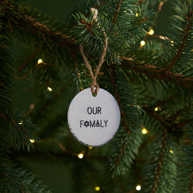 Może przedstawiać: Srebrna ozdoba choinkowa z rustykalnym sznurkowym zawieszeniem. Ozdoba jest okrągła i ma napisane na niej czarne litery "OUR FAMILY". Ozdoba wisi na zielonej choince.