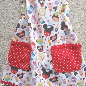 May include: White apron with a colorful pattern of cupcakes, donuts, and Mickey Mouse heads. Features two red polka dot pockets and red scalloped trim. The apron has a neck strap and waist ties.