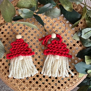 May include: Two handmade Christmas gnome ornaments. Each gnome has a red knitted hat, a white beard, and a wooden bead nose. The ornaments are hanging from a thin copper wire. The gnomes are displayed on a woven chair.