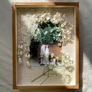 May include: A framed shadow box containing a wedding photograph of a couple kissing in front of a brick wall and greenery. The photo is surrounded by white lace, a pearl earring, and white baby's breath flowers.