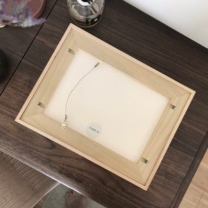 May include: The back of a light wood picture frame with a white mat and a wire hanging system. A small white sticker with the text "GAVE II" is attached to the frame.
