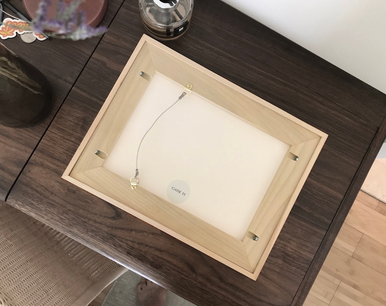 May include: The back of a light wood picture frame with a white mat and a wire hanging system. A small white sticker with the text "GAVE II" is attached to the frame.