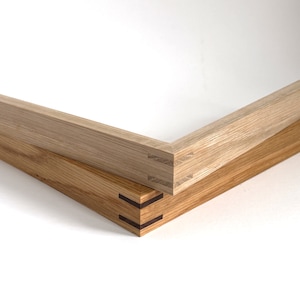 May include: Two wooden picture frames with mitered corners and a visible dovetail joint. The top frame is a lighter wood and the bottom frame is a darker wood.