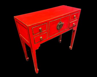Peut inclure: Une console en bois rouge avec trois tiroirs et une porte de cabinet avec une poignée en laiton. La table a une bordure dorée et repose sur quatre pieds.