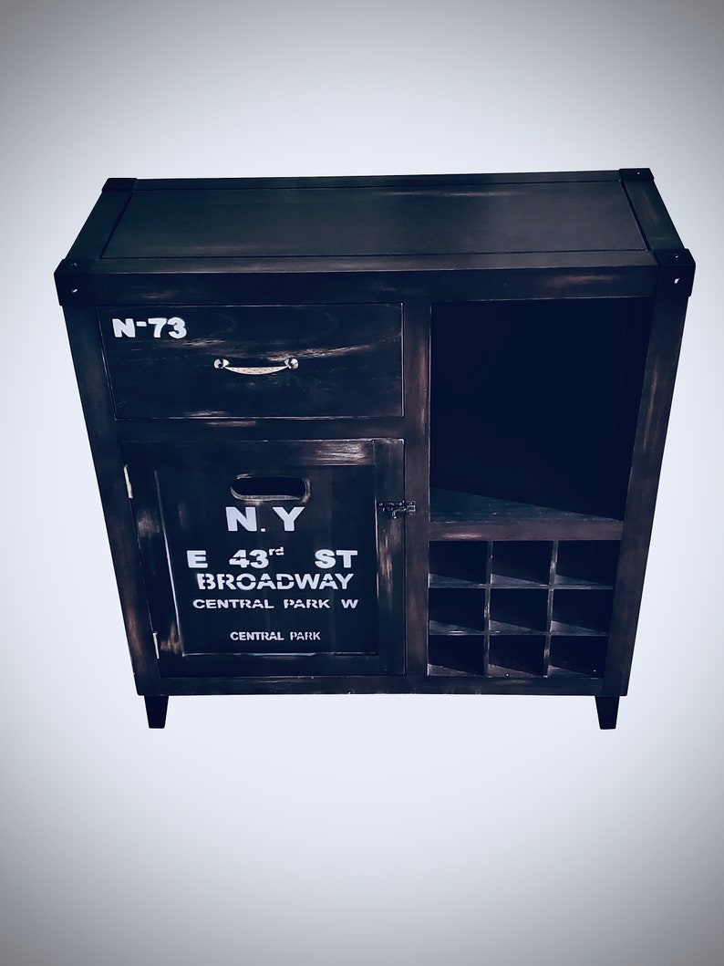 May include: Dark brown wooden cabinet with a drawer, door, and open shelf. The cabinet has the text "N-73" on the drawer and "N.Y. E 43rd ST BROADWAY CENTRAL PARK W CENTRAL PARK" on the door. The cabinet has a wine rack.
