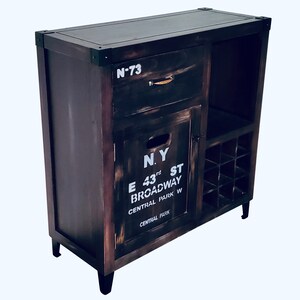 May include: A dark brown wooden cabinet with a distressed finish. It features a drawer, a door with the text "N.Y. E 43rd ST BROADWAY CENTRAL PARK W", and a wine rack. The cabinet has black metal corner accents and black legs.