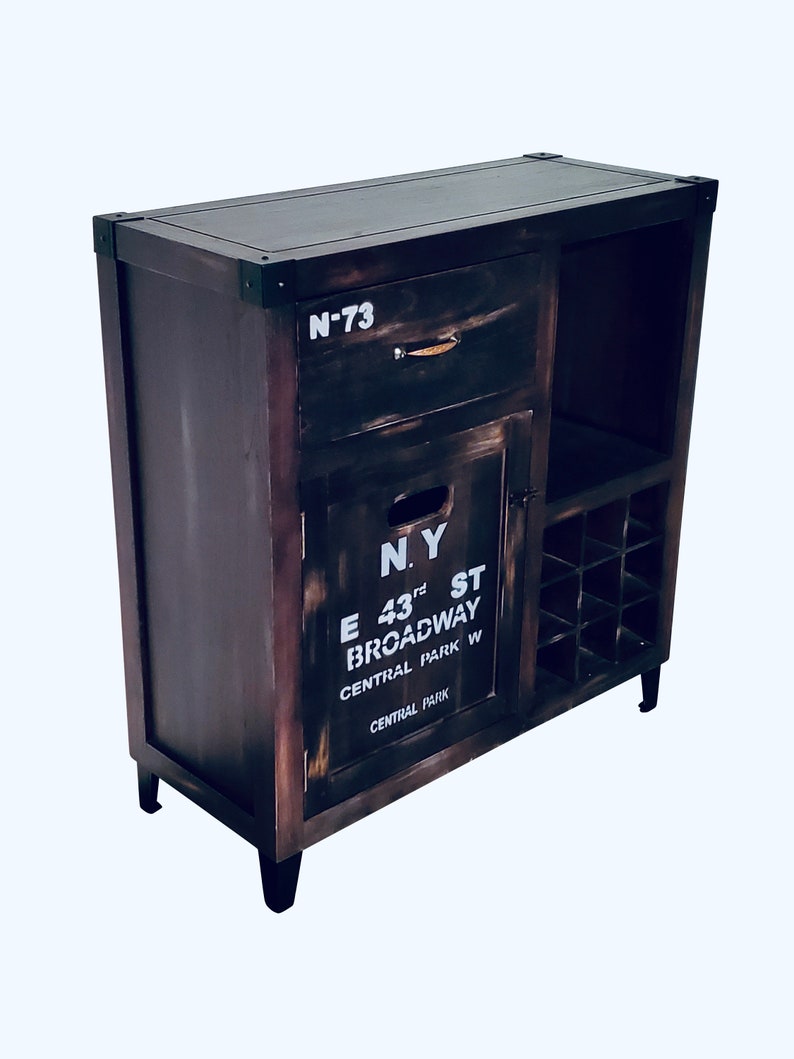 May include: A dark brown wooden cabinet with a distressed finish. It features a drawer, a door with the text "N.Y. E 43rd ST BROADWAY CENTRAL PARK W", and a wine rack. The cabinet has black metal corner accents and black legs.