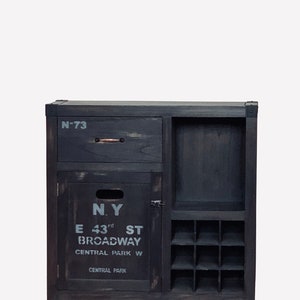 May include: Dark gray wooden cabinet with a drawer, door, and wine rack. The cabinet has the text "N.Y. E 43rd ST BROADWAY CENTRAL PARK W CENTRAL PARK" printed on the door. The drawer is labeled "N-73".