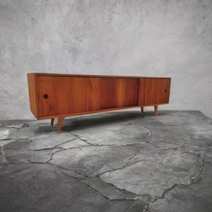 May include: A mid-century modern wooden cabinet with a warm brown finish. The cabinet has a long, rectangular shape with sliding doors and a pair of round handle holes. It stands on four angled, tapered legs, showcasing a minimalist design.