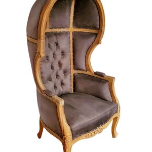 May include: A tall, wooden, hooded chair with a gray velvet upholstery. The chair has a rounded back and a carved wooden frame.