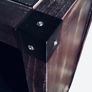 May include: Close-up of a dark brown wooden corner with a black metal corner bracket. The bracket is secured with three silver rivets. The wood grain is visible, and the overall aesthetic is rustic.