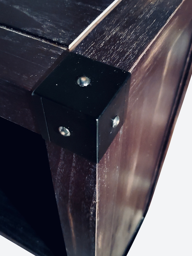 May include: Close-up of a dark brown wooden corner with a black metal corner bracket. The bracket is secured with three silver rivets. The wood grain is visible, and the overall aesthetic is rustic.