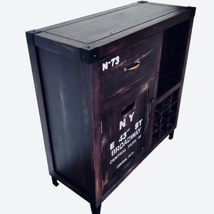 May include: Dark stained wooden bar cabinet with metal corner accents. Features a drawer, cabinet door with "N.Y. E 43rd St Broadway Central Park W" text, and a wine rack. The cabinet has a distressed finish.