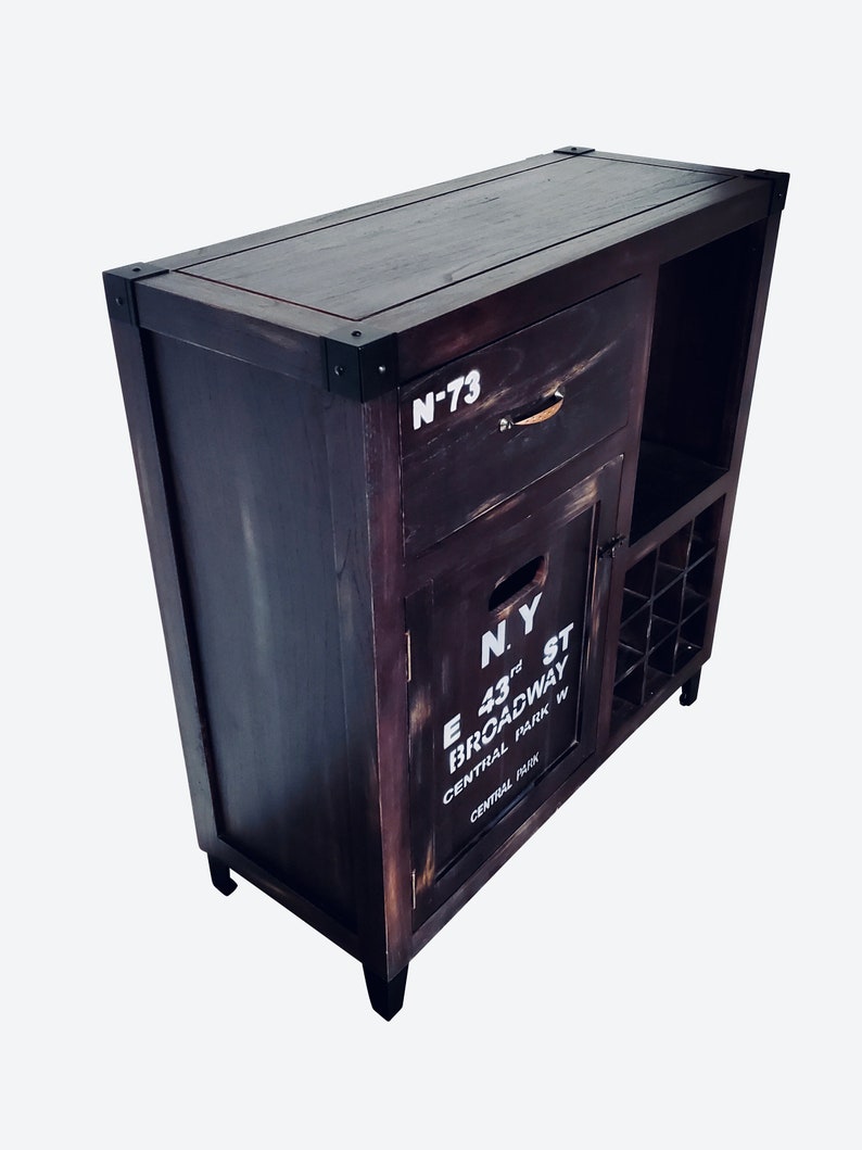 May include: Dark stained wooden bar cabinet with metal corner accents. Features a drawer, cabinet door with "N.Y. E 43rd St Broadway Central Park W" text, and a wine rack. The cabinet has a distressed finish.