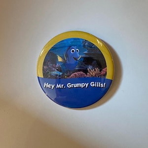 May include: A circular pinback button featuring a blue fish character swimming in an underwater coral reef scene. The bottom section is dark blue with the white text "Hey Mr. Grumpy Gills!" printed in a bold, sans-serif font against a yellow border.