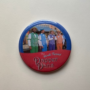 May include: A round button with a blue border and a red lower section. The button features a picture of four men in striped vests and straw hats. The text "The World Famous Dapper Dans" is printed on the button.