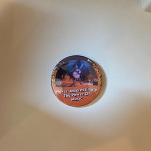 May include: A round button with a musical theme. The button features an illustration of a musician playing a guitar, surrounded by autumn leaves. The text "Never Underestimate The Power Of Music" is printed on the button.