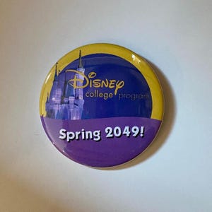 May include: A circular pinback button featuring a yellow border, a blue center with a graphic of a castle, and the text Disney college program. The bottom section is purple with the white text Spring 2049! printed in a bold, sans-serif font.