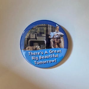 May include: A round, blue button with the text "There's A Great Big Beautiful Tomorrow!" in white. The button features a vintage illustration of a man, a dog, and a room interior.
