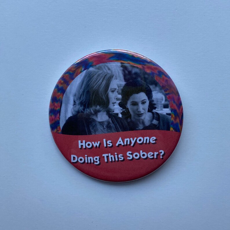 May include: A round button with a red base and a colorful psychedelic border. The button features a black and white image of two women and the text "How Is Anyone Doing This Sober?"