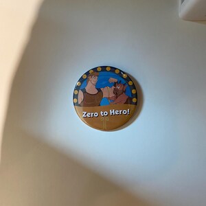 May include: A round button featuring a cartoon image of Hercules and a creature. The button has a gold border with blue dots and the text "Zero to Hero!" in white. The background is a light tan color.