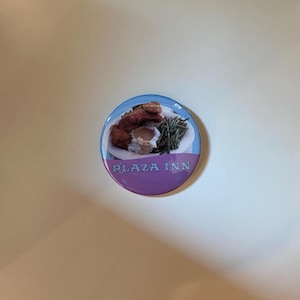May include: A round button featuring a plate of fried chicken, mashed potatoes, green beans, and gravy. The button has a blue and purple border with the text "PLAZA INN".