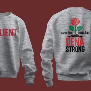 以下が含まれることがあります： 前面に赤い文字で「RESILIENT」と印刷され、背面に「ASHES FADE ROSES RISE DENA STRONG」というテキストと赤いバラと緑の葉が印刷された灰色のスウェットシャツです。