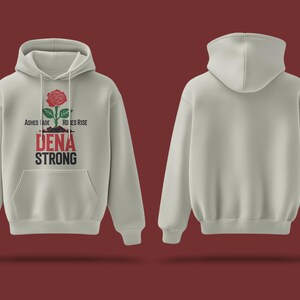 K&ouml;nnte beinhalten: Ein hellgrauer Hoodie mit einem roten Rosenmotiv und dem Text "Ashes Made Roses Rise Dena Strong". Der Hoodie hat eine Kapuze mit Kordelzug und eine K&auml;ngurutasche.