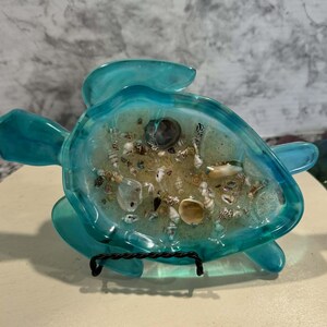 May include: A turquoise resin turtle-shaped dish filled with sand and seashells. The dish is supported by a black metal stand. The turtle's flippers and head extend outward, creating a decorative coastal theme.