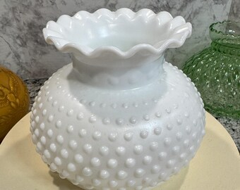 May include: A white hobnail glass vase with a ruffled rim. The vase has a round body covered in small, raised bumps. The vase sits on a light yellow surface, with a blurred background.