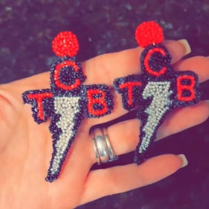 Puede incluir: Un par de pendientes de cuentas. Cada pendiente presenta una parte superior de cuentas rojas, las letras "TCB" en rojo y un rayo plateado. Los pendientes son negros y plateados con un acento rojo.