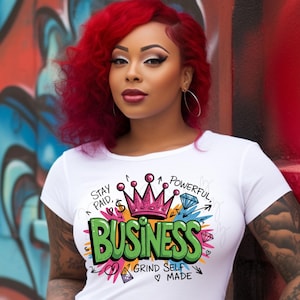May include: White t-shirt with a colorful graphic that says "BUSINESS" in green, with a pink crown and other design elements. The shirt also has the phrases "Stay Paid, Powerful, Grind Self Made". The model is wearing blue jeans.