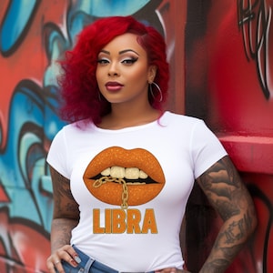 May include: White t-shirt featuring a graphic of orange lips with a gold chain and the word "LIBRA" in orange. The shirt is paired with blue jeans. The background is a graffiti wall with red, blue, and black paint.